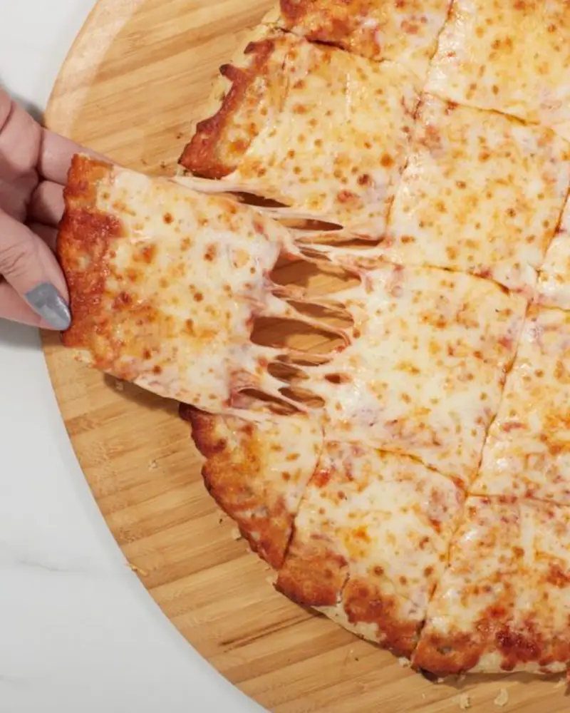 An overhead close-up of a square-cut Chicago tavern-style cheese pizza on a round wooden cutting board. A hand with silver nail polish is pulling away a crispy edge piece, creating a stretched, gooey cheese pull.