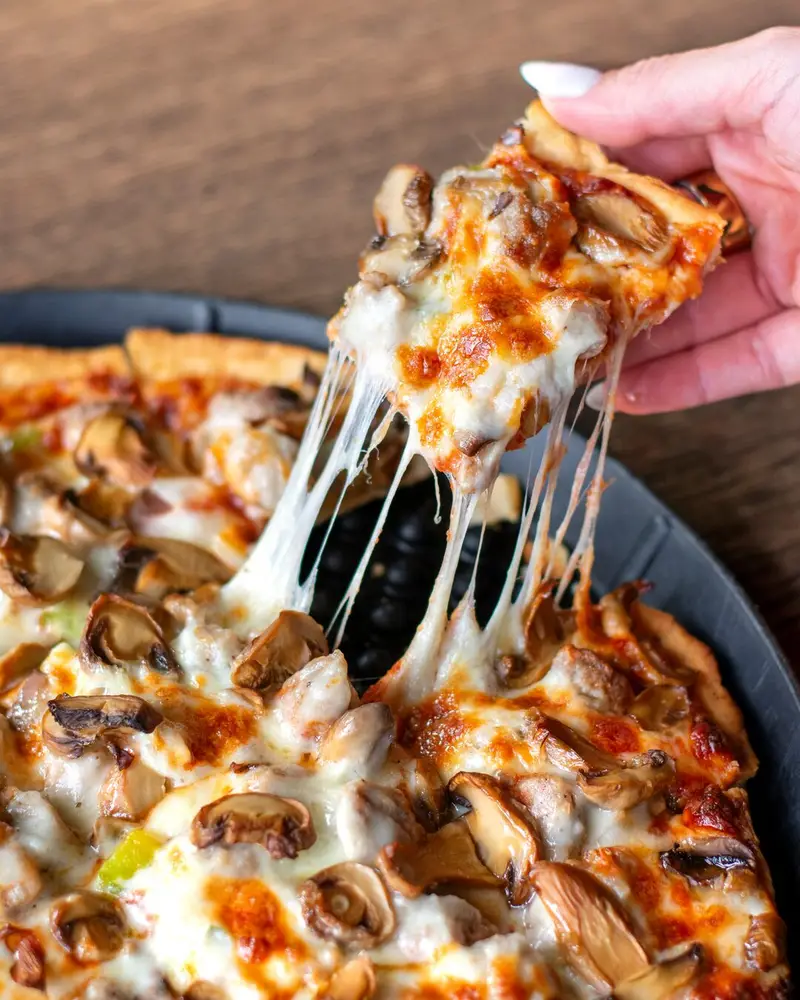 A close-up shot of a hand lifting a slice of thick-crust pizza loaded with melted mozzarella cheese, sliced mushrooms, and sausage, creating a dramatic, stretchy cheese pull from the pie below.
