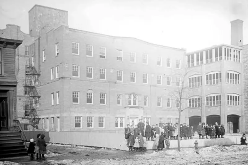 A historical black-and-white photograph showing a large group of immigrant children and adults standing outside a brick Hull-House settlement building in the winter.