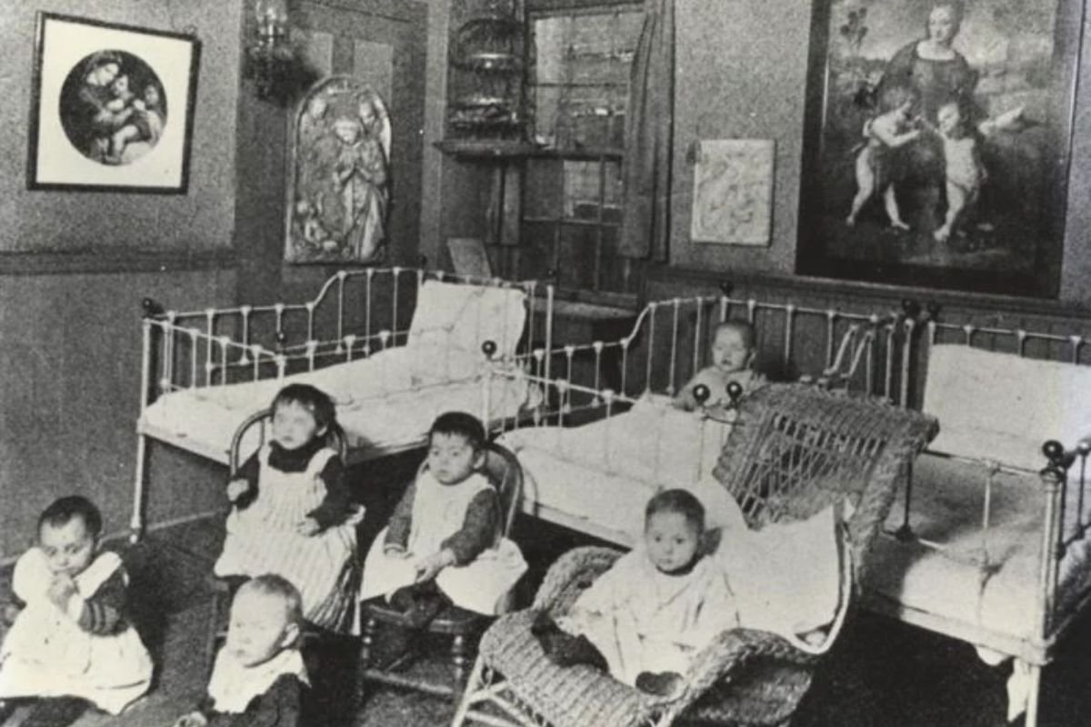 A historical black-and-white photograph from the 1890s showing toddlers sitting in wicker chairs and iron cribs inside the Hull-House nursery in Chicago.