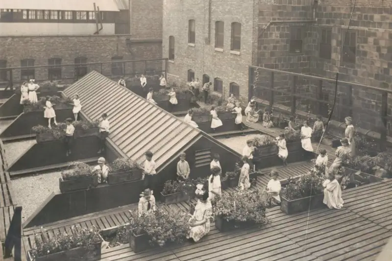 A 1912 historical black-and-white photograph showing immigrant children tending to plants on the rooftop garden of the Elizabeth McCormick Open Air School at Chicago's Hull-House.