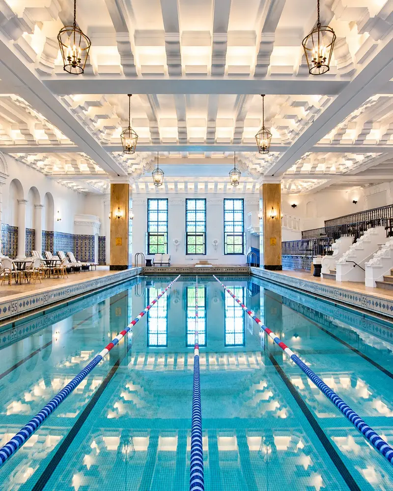 A long, pristine indoor lap pool stretches into the distance under an ornate, bright white coffered ceiling illuminated by hanging lantern lights. The calm turquoise water reflects the row of large windows at the far end, while multi-tiered spectator seating and decorative white columns line the right side of the grand room.
