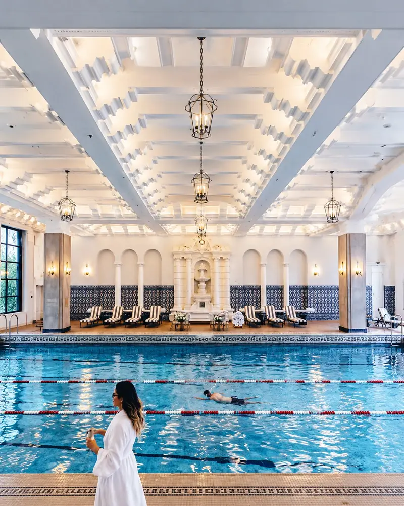 A large, luxurious indoor swimming pool features clear blue water divided by lane lines, set beneath an ornate white coffered ceiling with hanging lantern chandeliers. A swimmer moves through the water while a person in a white robe stands in the foreground, and the background shows a row of lounge chairs against decorative tiled walls.