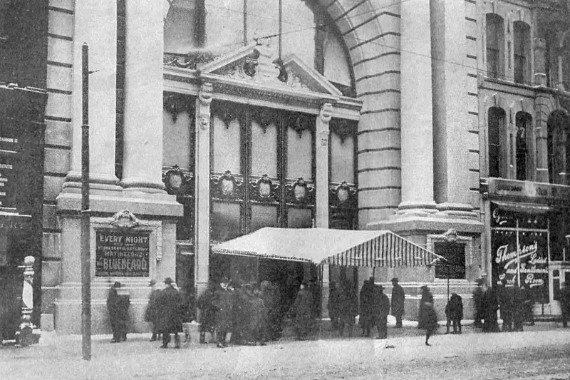 Archival black-and-white photograph of the exterior of the Iroquois Theatre in Chicago, showing a crowd gathered under an awning and a stone marquee advertising the play Bluebeard.