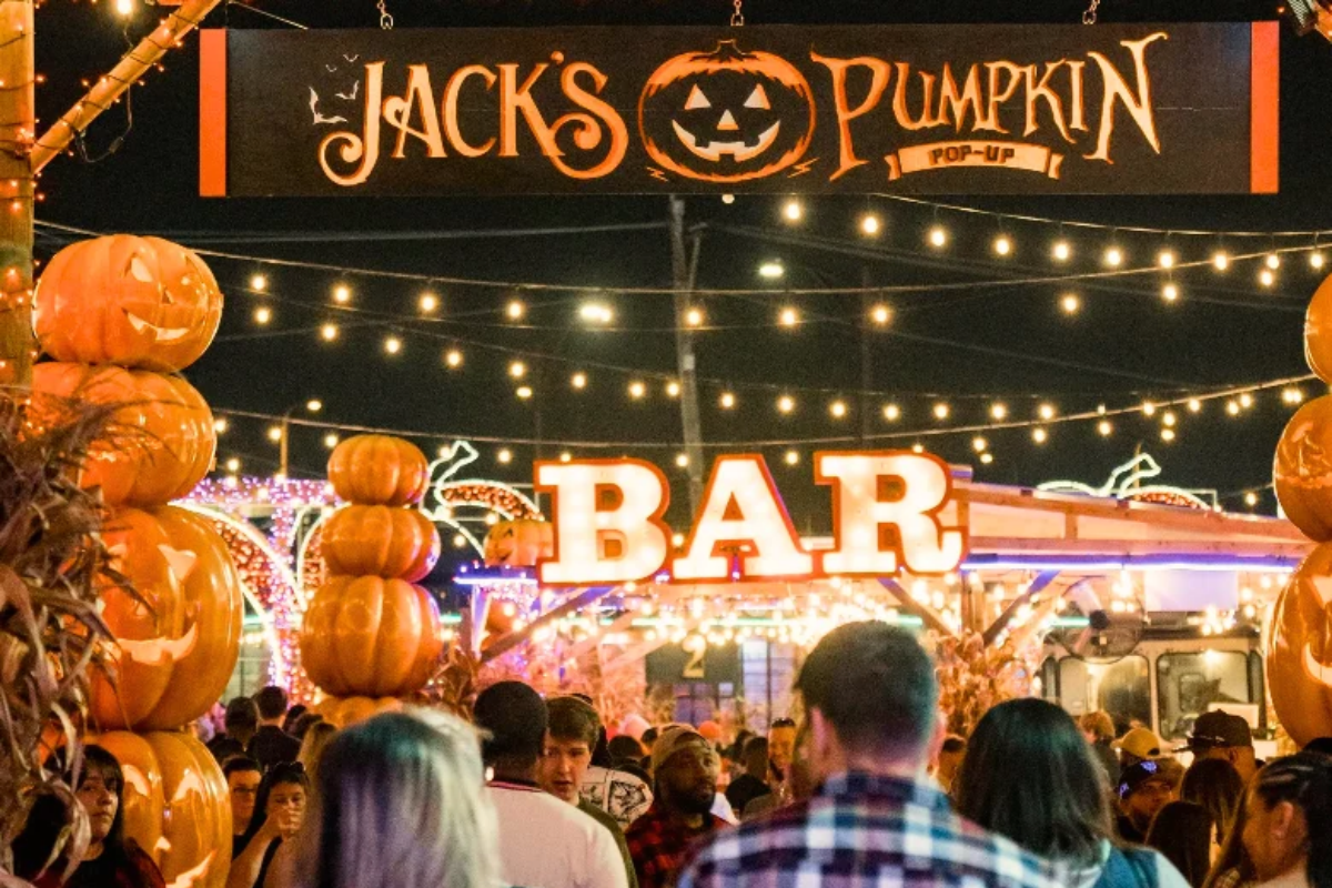 A bustling nighttime crowd walks under glowing string lights and a large marquee "BAR" sign at Jack's Pumpkin Pop-Up, flanked by tall columns of glowing orange pumpkins.