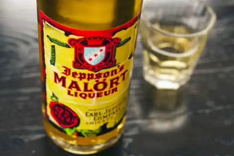 A close-up photograph of a Jeppson's Malört bottle featuring its iconic yellow and red three-star label, resting on a dark wooden bar top next to a poured shot glass.