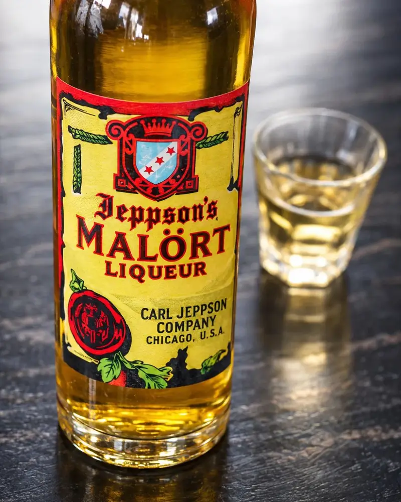 A bottle of Jeppson's Malört liqueur next to a poured shot glass resting on a dark countertop.