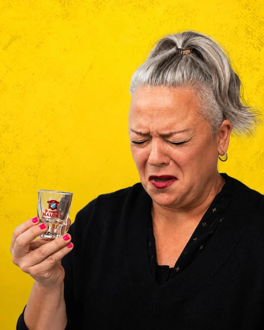 A woman with grey hair tied in a ponytail winces in a dramatic "Malört face" reaction after taking a shot from a Jeppson's Malört glass. She tightly shuts her eyes and grimaces with a look of distaste against a bright, textured yellow background.