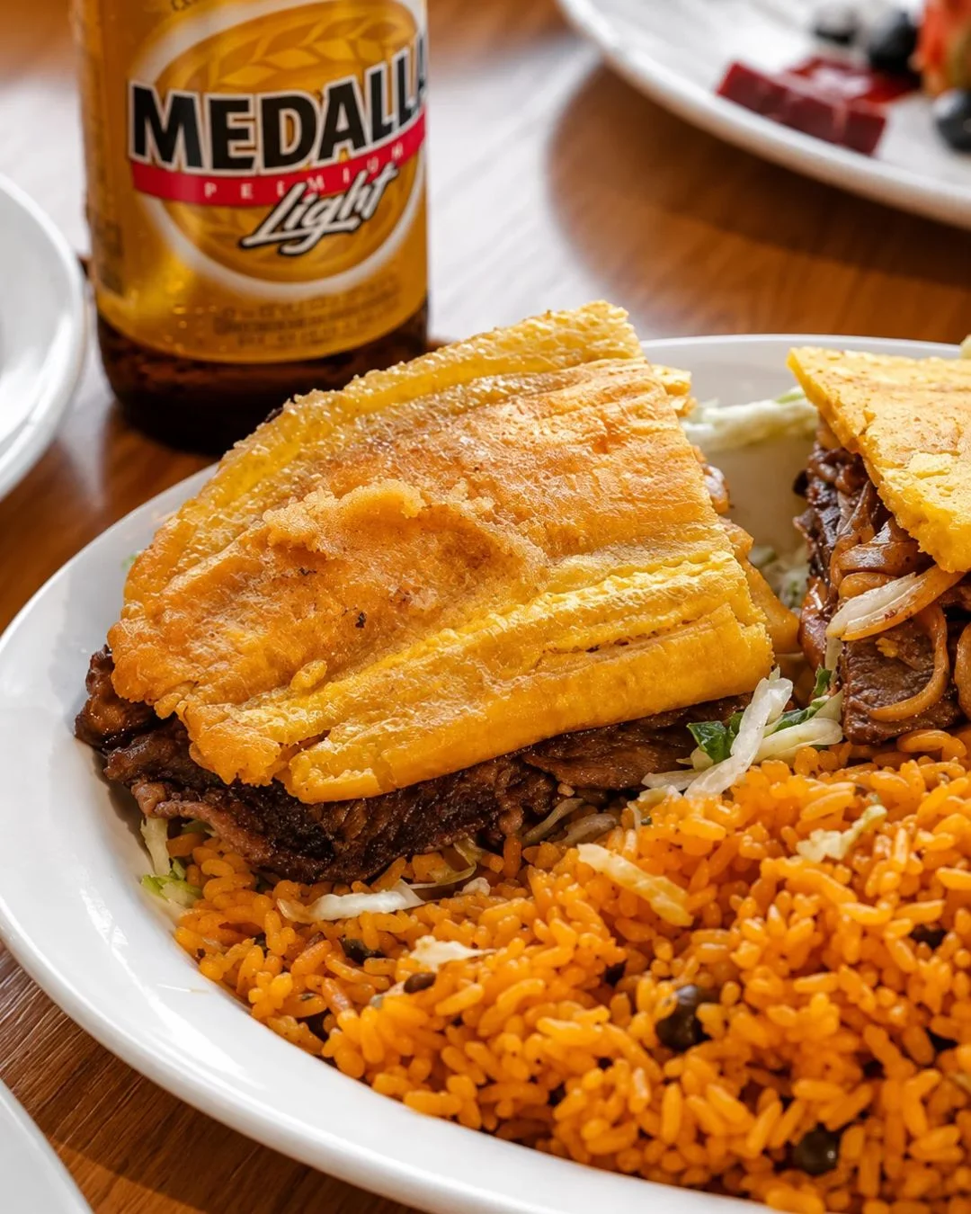 A close-up view of a Puerto Rican jibarito sandwich made with crispy fried green plantains instead of bread, filled with seasoned steak, lettuce, and onions, served on a white plate with a side of bright orange Puerto Rican rice next to a bottle of Medalla Light beer.