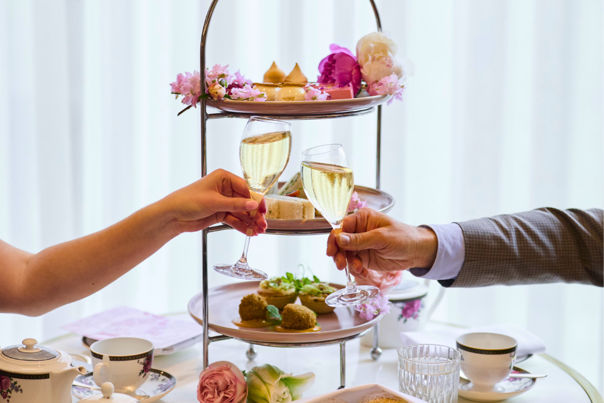 Two people clinking glasses of champagne in front of a three-tiered silver stand filled with gourmet pastries, finger sandwiches, and flowers during a luxury afternoon tea service.