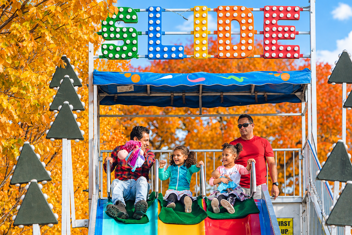 A family smiling as they ride down a colorful carnival slide surrounded by bright yellow autumn foliage at a fall festival.