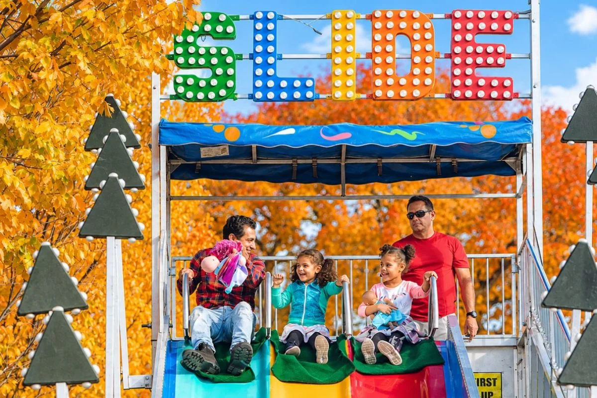 A family with two young girls smiling as they ride down a colorful carnival slide under a bright, multi-colored "SLIDE" sign, surrounded by vibrant yellow and orange autumn trees at a Chicago fall festival.