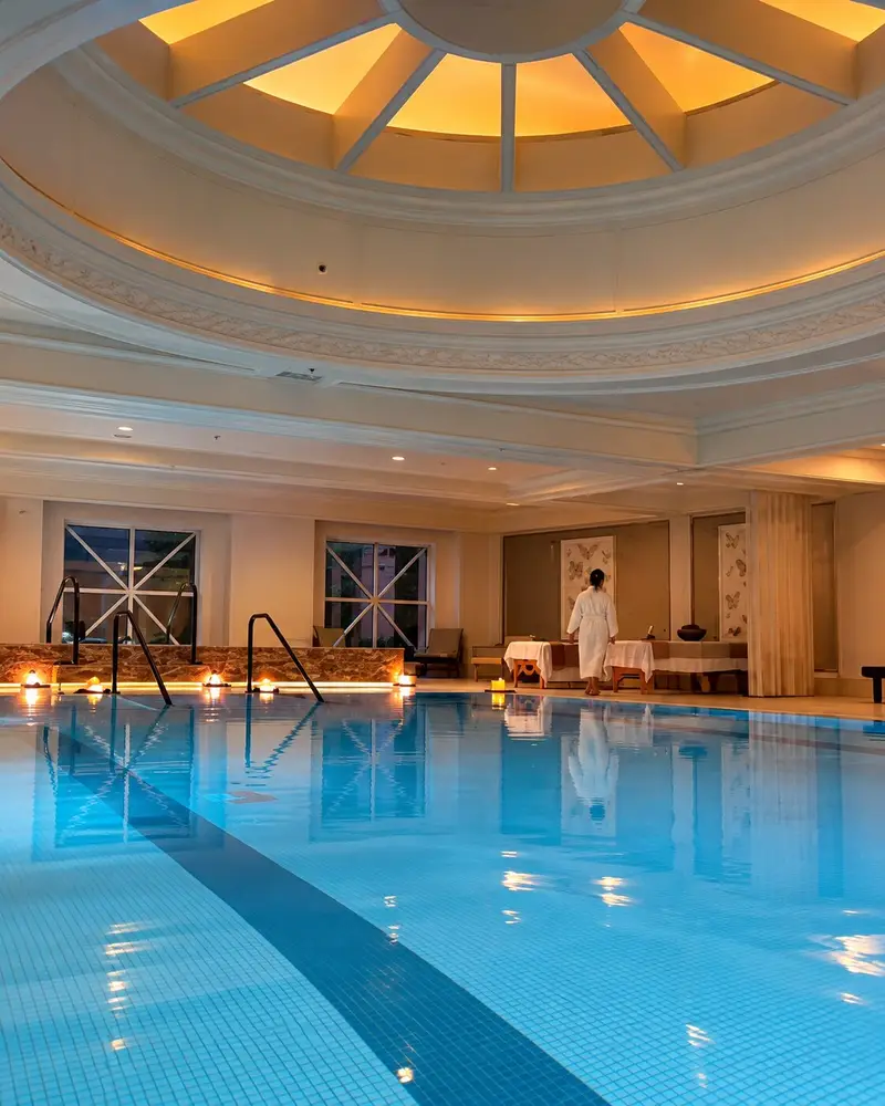 A calm, blue indoor swimming pool reflects a large, illuminated circular skylight dome embedded in an elegant white ceiling. In the background, a person in a white bathrobe stands near spa tables in a softly lit relaxation area flanked by windows and candlelight accents.