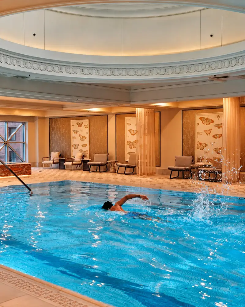 A swimmer glides through the clear turquoise water of an elegant indoor pool, leaving a splash behind them. The Mediterranean-style pool room is adorned with an ornate domed ceiling and cream-colored columns, while warm lighting illuminates comfortable lounge chairs and butterfly-themed art panels along the back wall.