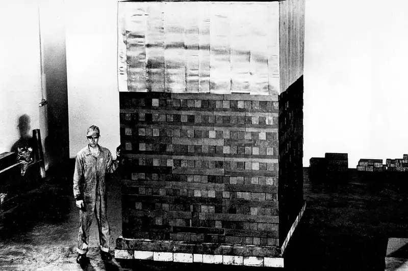 Black and white archival photograph of a Manhattan Project worker in coveralls standing next to a massive, square stack of graphite blocks used in early nuclear test piles at the University of Chicago.