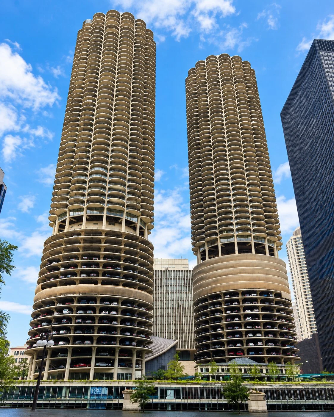 Two identical, cylindrical concrete skyscrapers with unique corncob-like designs rise dramatically against a partly cloudy sky. The lower tiers of the iconic buildings feature open-air, spiral parking decks filled with visible cars.