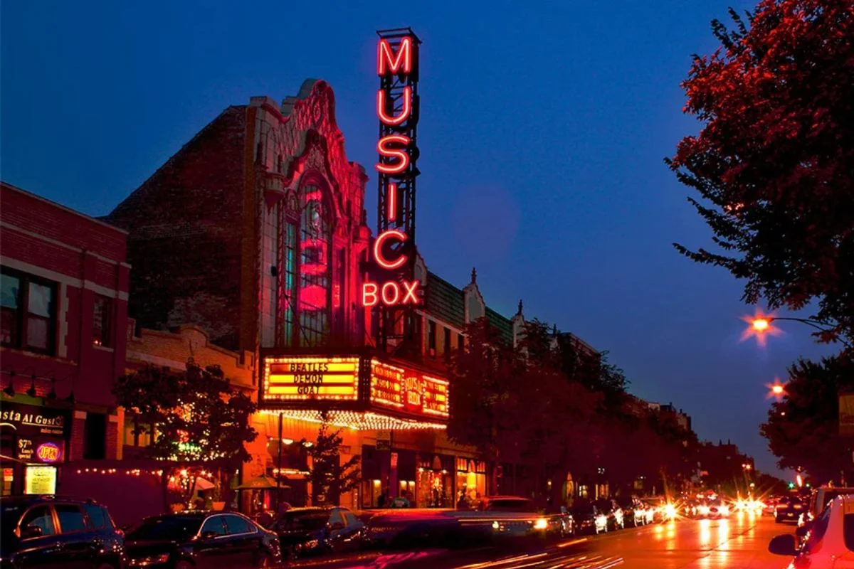 The iconic red neon Music Box Theatre sign and bright marquee glowing against a twilight sky above a busy Chicago street lined with cars and streetlights.