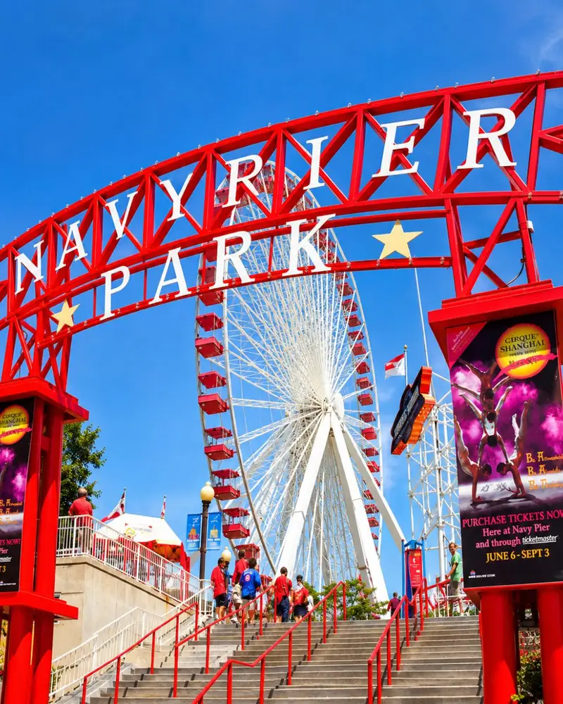 A large, bright red metal archway displaying the white text "NAVY PIER PARK" frames a massive white Centennial Wheel with red passenger gondolas under a clear blue sky. Concrete steps with red railings lead up toward the entrance, where several visitors are walking up to the park. Promotional banners for a Cirque Shanghai show flank both sides of the entrance structure.