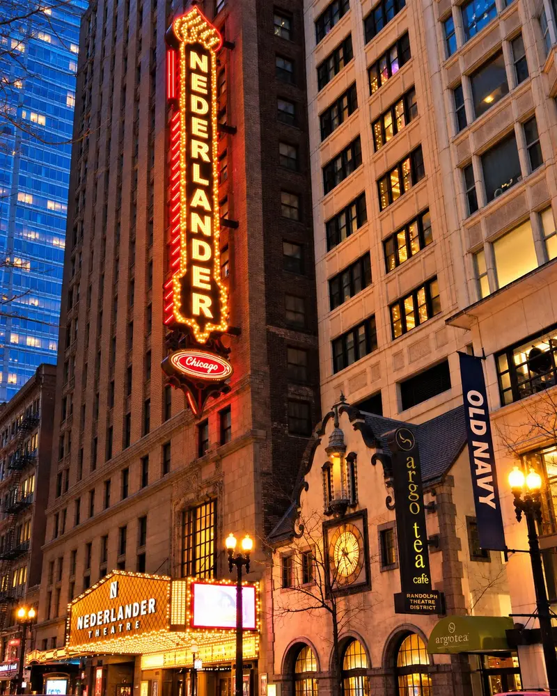 A tall, vertical marquee sign reading "NEDERLANDER" glows with bright orange and white neon letters on the side of a historic stone skyscraper at dusk. Below it, an illuminated street-level canopy marquee for the Nederlander Theatre stands adjacent to a classic European-style building featuring a prominent lit clock face .