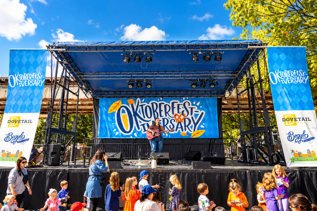 An outdoor festival stage with a large blue Oktoberfestiversary banner featuring an acoustic musician performing for a crowd of families and young kids under a clear sky at the Begyle and Dovetail Brewery street party in Chicago.