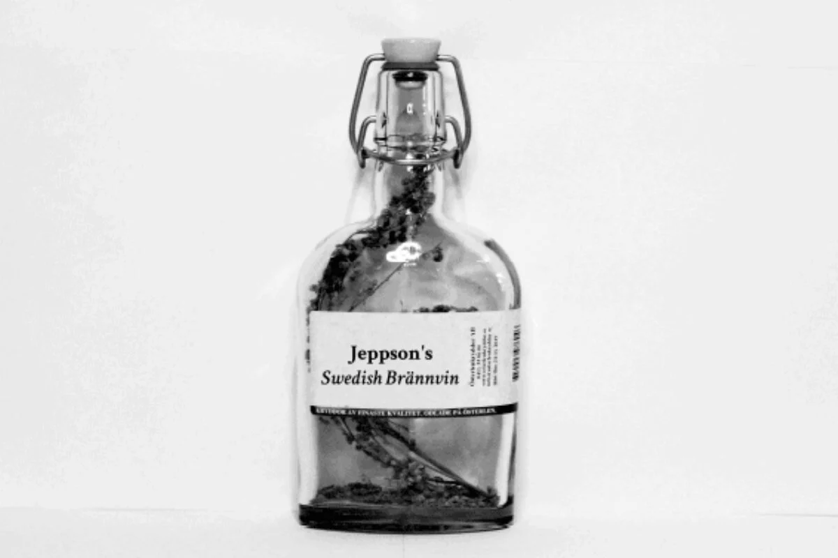 A black and white photograph of a glass swing-top bottle labeled Jeppson's Swedish Brännvin, featuring raw sprigs of wormwood steeping inside the clear liquor.
