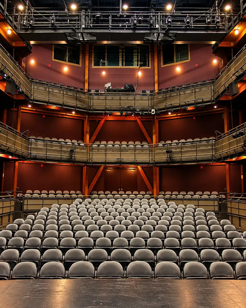 An empty courtyard-style auditorium features neatly arranged rows of curved white seats on the main floor facing a dark stage setup. Above, two levels of matching curved balcony seating options wrap around the intimate space under overhead production rigging.