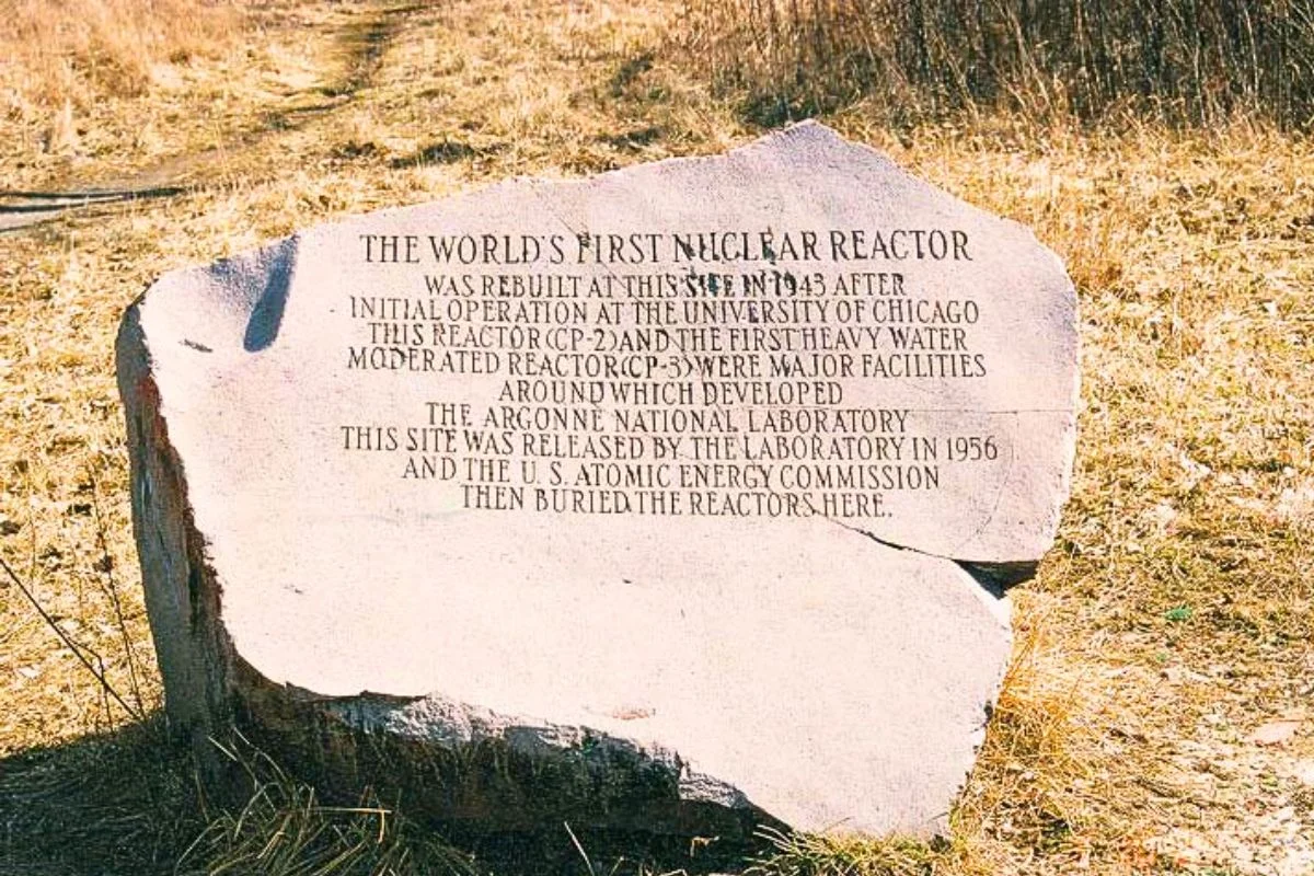 Photograph of a large, rough-hewn stone monument sitting in a grassy clearing in Red Gate Woods. The engraved text explains that the world's first nuclear reactor from the University of Chicago was rebuilt and eventually buried at this exact site by the U.S. Atomic Energy Commission.