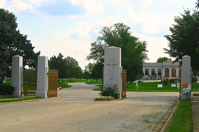 The stone gates and entrance pillars of Resurrection Cemetery in Justice, Illinois, showing a carved cross, open iron gates, and a paved driveway.