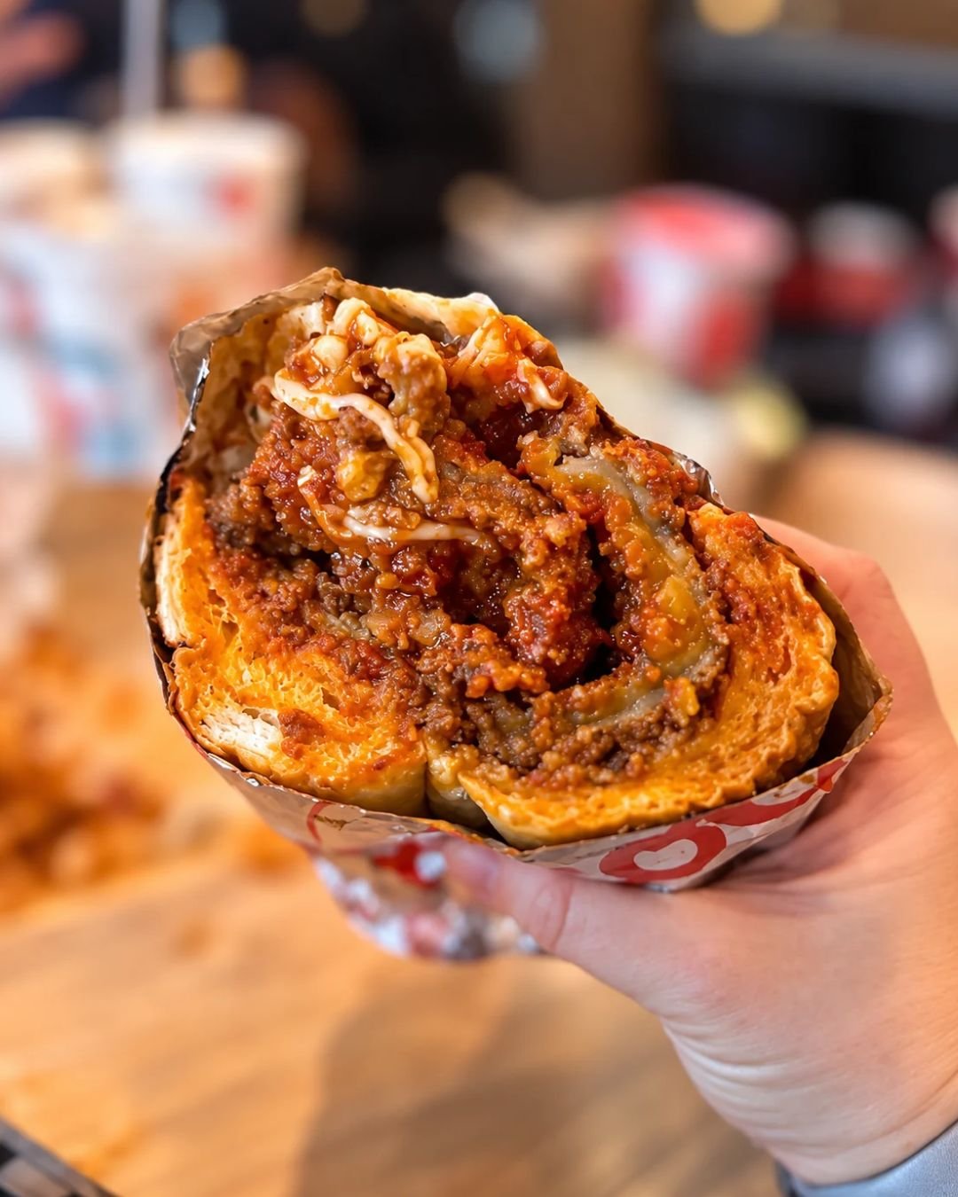 A close-up, first-person view of a hand holding a cross-section of a famous Chicago breaded steak sandwich wrapped in foil. The thick Italian roll is packed with crispy breaded steak, rich red marinara meat sauce, and melted mozzarella cheese.