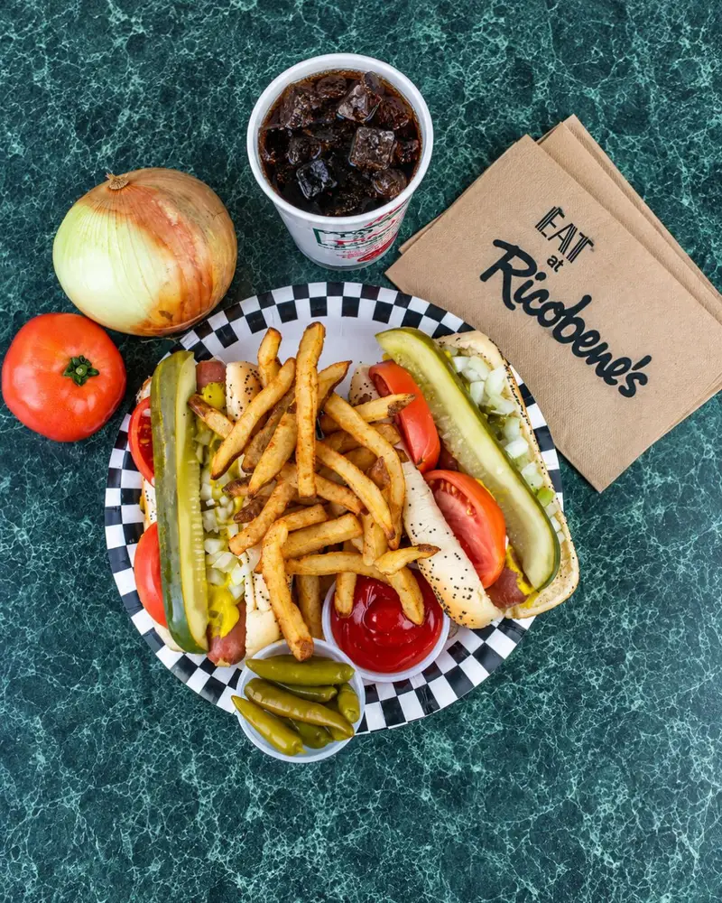 An overhead flat-lay view of two Chicago-style hot dogs loaded with pickle spears, tomatoes, and onions on a checkered plate, piled with french fries, served with a soda, small peppers, ketchup, a whole tomato, an onion, and Ricobene's branded napkins on a green marbled table.