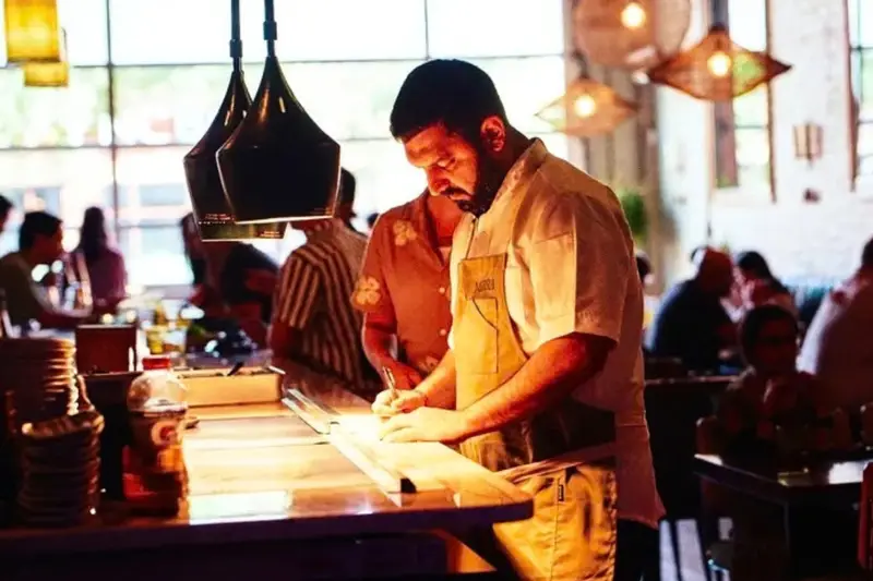 Chef Rishi Kumar writing at the illuminated open kitchen counter during a busy dinner service at Mirra restaurant in Bucktown, Chicago.
