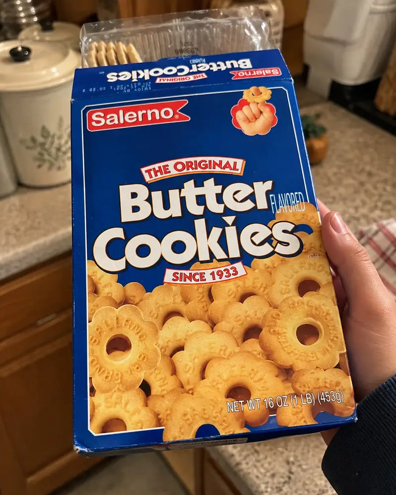 A hand holds up a classic blue box of Salerno Original Butter Cookies on a kitchen countertop background. The vintage-style packaging prominently features a bold white title, a graphic of stacked flower-shaped cookies with holes in the center, and a small illustration of a child's hand holding up a cookie.
