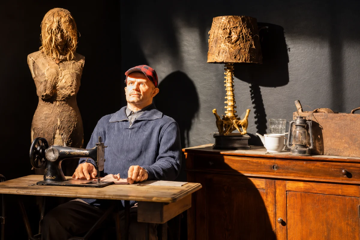 A life-sized museum diorama at the Serial Killer Exhibition in Chicago featuring a lifelike figure of Ed Gein sitting at a vintage sewing machine, next to a macabre lamp made of bones.