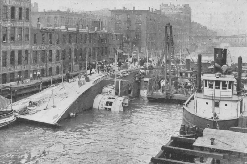 Archival black-and-white photograph of the SS Eastland capsized on its side in the Chicago River. Rescue workers and survivors stand on the exposed white hull just feet from the city docks, with a tugboat assisting in the foreground.