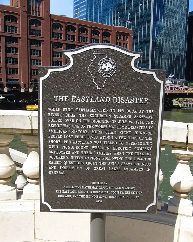 A close-up of the bronze Illinois State Historical Society memorial plaque for The Eastland Disaster located on the Chicago Riverwalk, with the historic red-brick Reid, Murdoch & Co. building visible across the river in the background.