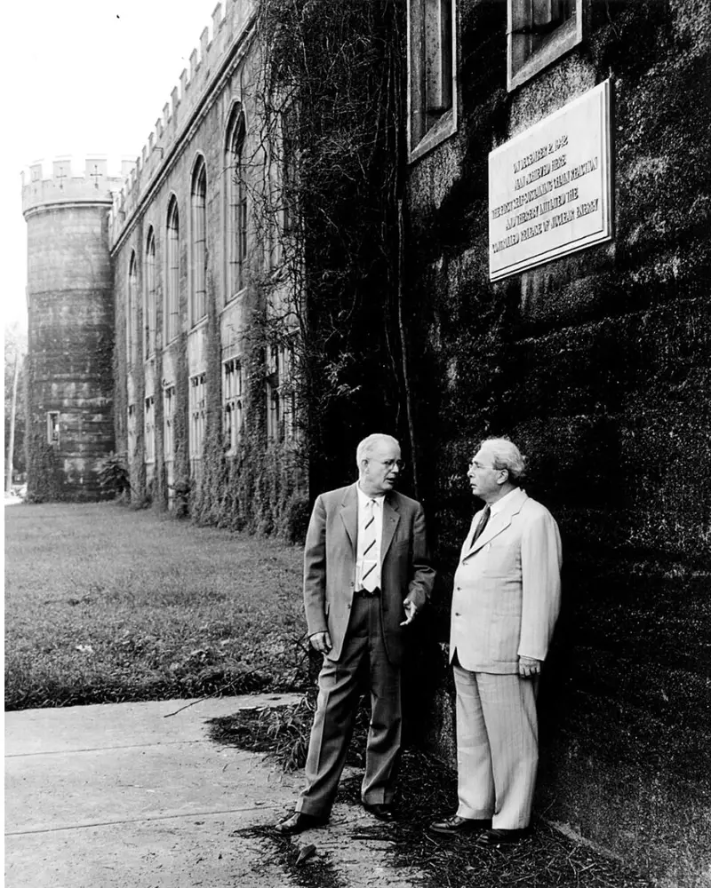Black and white archival photograph of Manhattan Project scientists Norman Hilberry and Leo Szilard standing outside the ivy-covered stone exterior of Alonzo Stagg Field beneath a bronze plaque commemorating the first nuclear chain reaction.