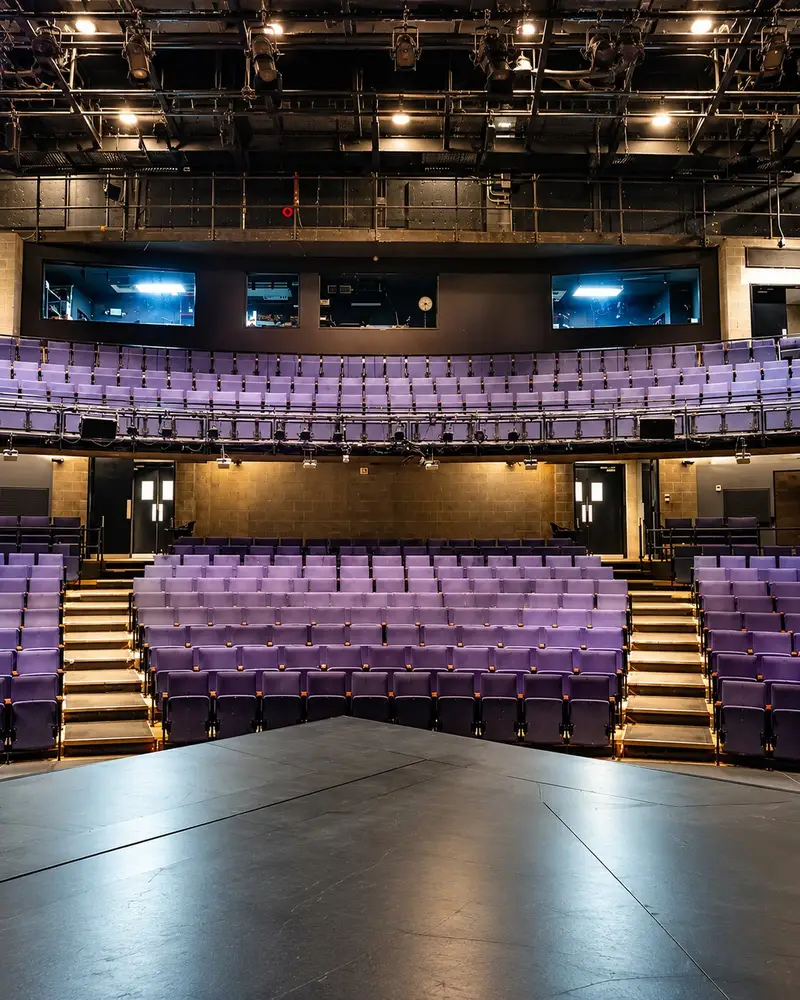 An empty auditorium features rows of purple velvet seats neatly tiered on steps leading up to an illuminated back wall. The shot is taken from a dark stage platform looking toward the curved seating configuration and upper control booths.