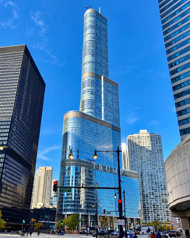 The massive, blue-glass Trump International Hotel and Tower rises high into a clear sky, featuring distinct asymmetric setbacks as it tapers upward. In the foreground, city streetlights, a Wacker Drive street sign, and pedestrians frame the base of the gleaming skyscraper.