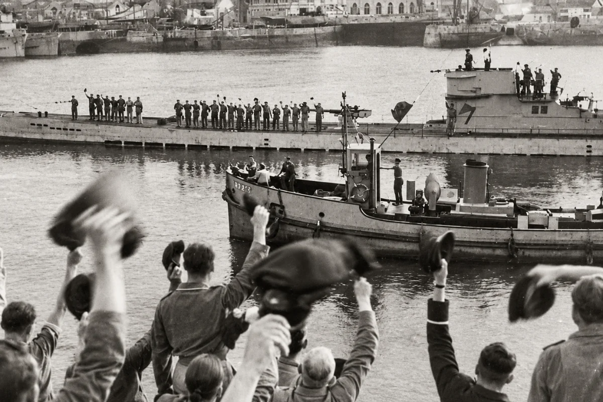 Black and white historical photograph of a German U-boat crew waving from the deck of their submarine in a harbor, with onlookers in the foreground cheering and waving hats in the air.