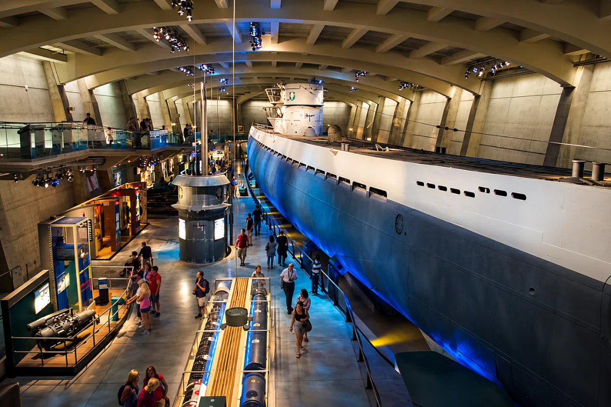A wide, brightly lit view of the massive U-505 submarine displayed inside a modern, concrete underground bunker at the Griffin Museum of Science and Industry in Chicago, featuring dramatic blue lighting and museum visitors walking alongside the hull.