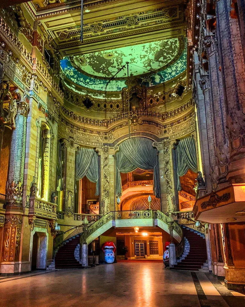 The ornate grand lobby of the historic Uptown Theatre in Chicago, featuring a sweeping double staircase, heavy drapes, gold architectural details, and peeling paint on a domed ceiling.