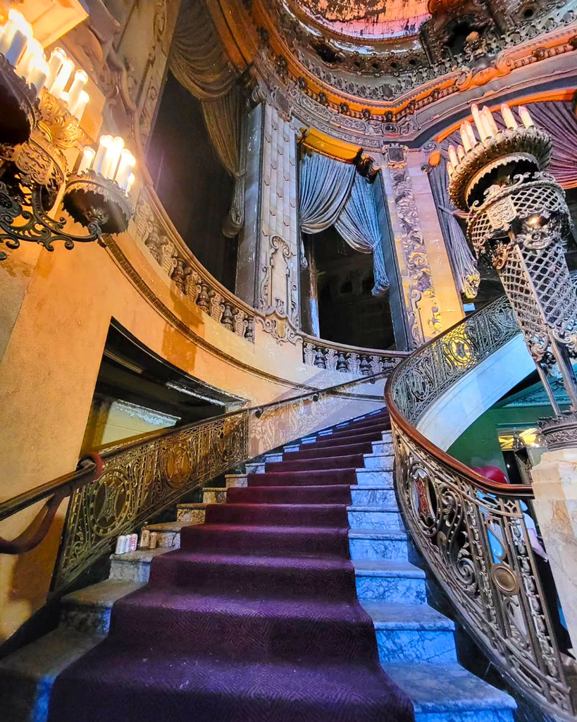A low-angle view of the grand marble staircase inside the historic Uptown Theatre in Chicago, featuring a red carpet, ornate metal railings, and decaying Spanish Baroque Revival architectural details.