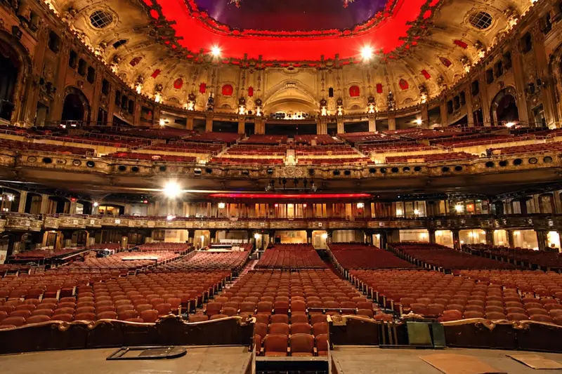 The massive 4,400-seat auditorium of the historic Uptown Theatre in Chicago, featuring ornate Spanish Baroque Revival architecture and rows of empty seats under a red illuminated ceiling.