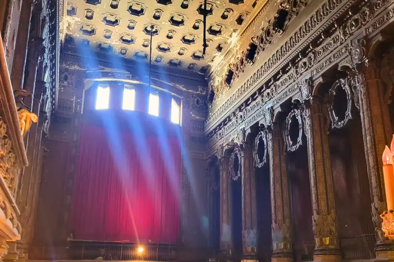 Dramatic sunbeams shine through high windows into the dusty interior of the historic Uptown Theatre in Chicago, illuminating a red stage curtain and a decaying, ornate coffered ceiling.
