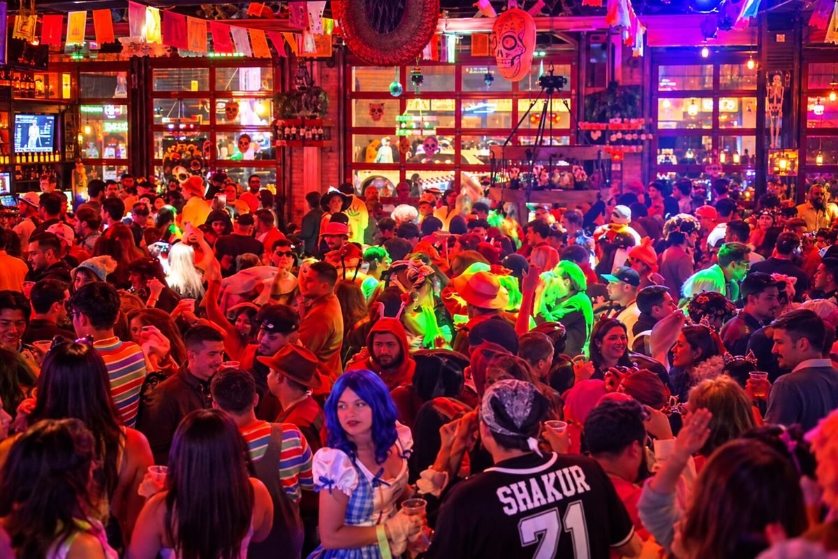 A packed, indoor bar venue is filled to capacity with a dense crowd of young adults wearing colorful Halloween costumes. Bright, festive neon lighting in shades of pink, orange, and green illuminates the lively atmosphere beneath hanging Dia de los Muertos banners.