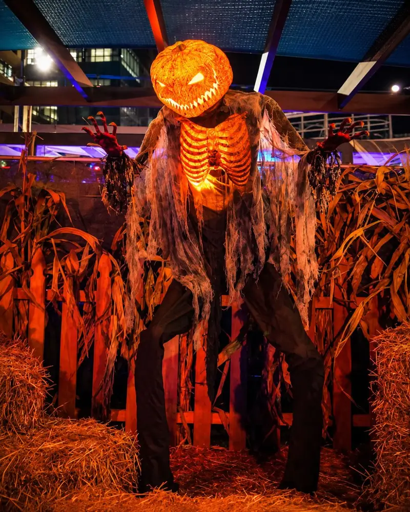 A towering, menacing Halloween prop features a glowing, fiercely grinning pumpkin head and a lit-up orange rib cage. The creature is draped in tattered grey cloth and stands in a spooky autumn display surrounded by a wooden fence, corn stalks, and hay bales.